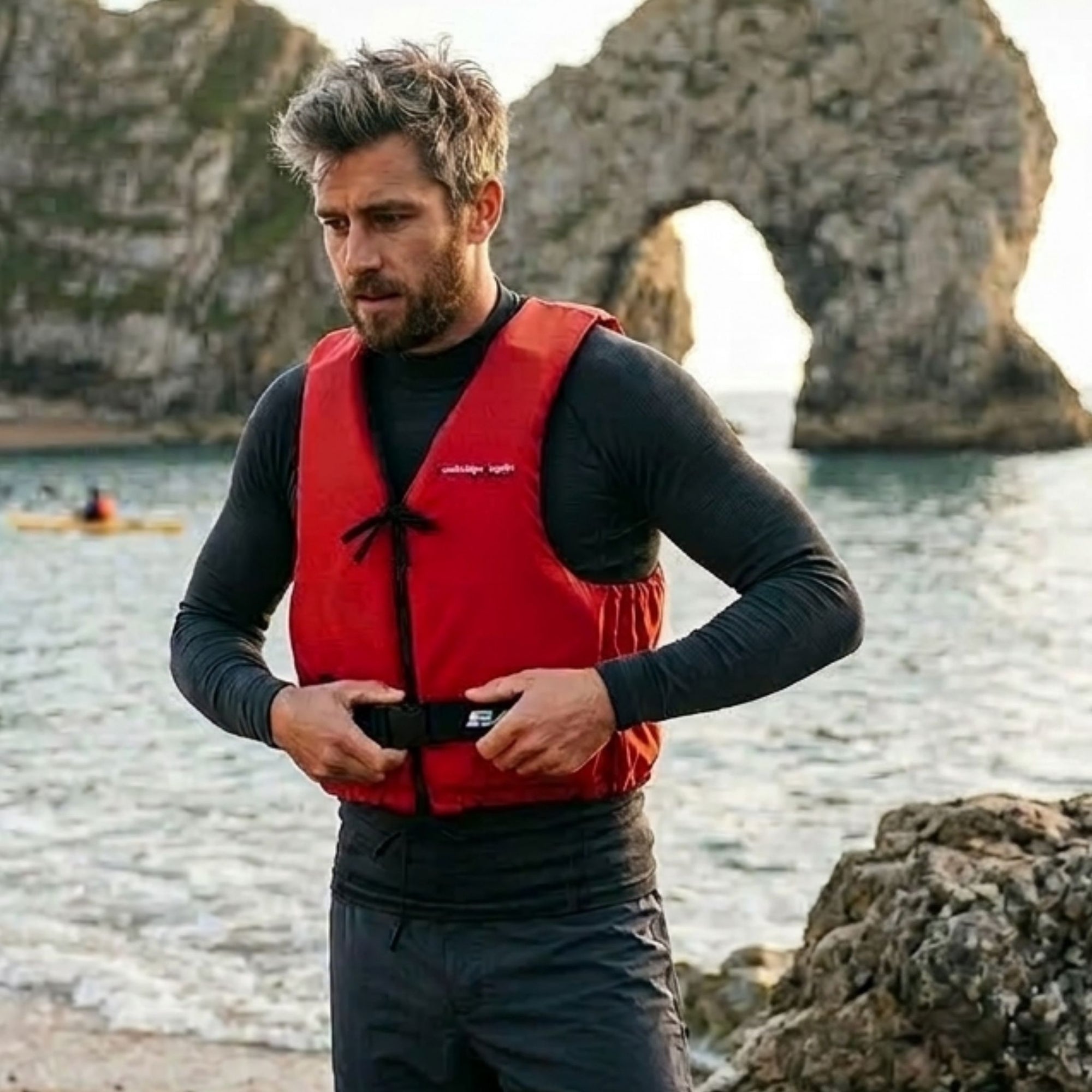 Man wearing Baltic Aqua Pro red canoe buoyancy aid adjusting the waist strap while standing on a rocky beach with sea arch cliffs behind, designed for kayaking, canoeing, and paddle sports safety.
