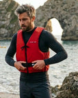 Man wearing Baltic Aqua Pro red canoe buoyancy aid adjusting the waist strap while standing on a rocky beach with sea arch cliffs behind, designed for kayaking, canoeing, and paddle sports safety.