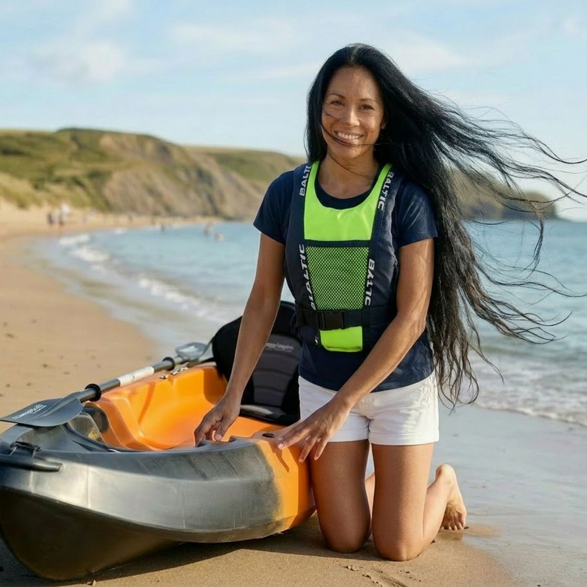 Woman kneeling on a sandy beach beside a sit-on-top kayak wearing a Baltic canoe buoyancy aid with green mesh front panel and adjustable waist belt.