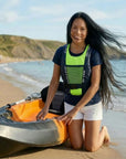 Woman kneeling on a sandy beach beside a sit-on-top kayak wearing a Baltic canoe buoyancy aid with green mesh front panel and adjustable waist belt.