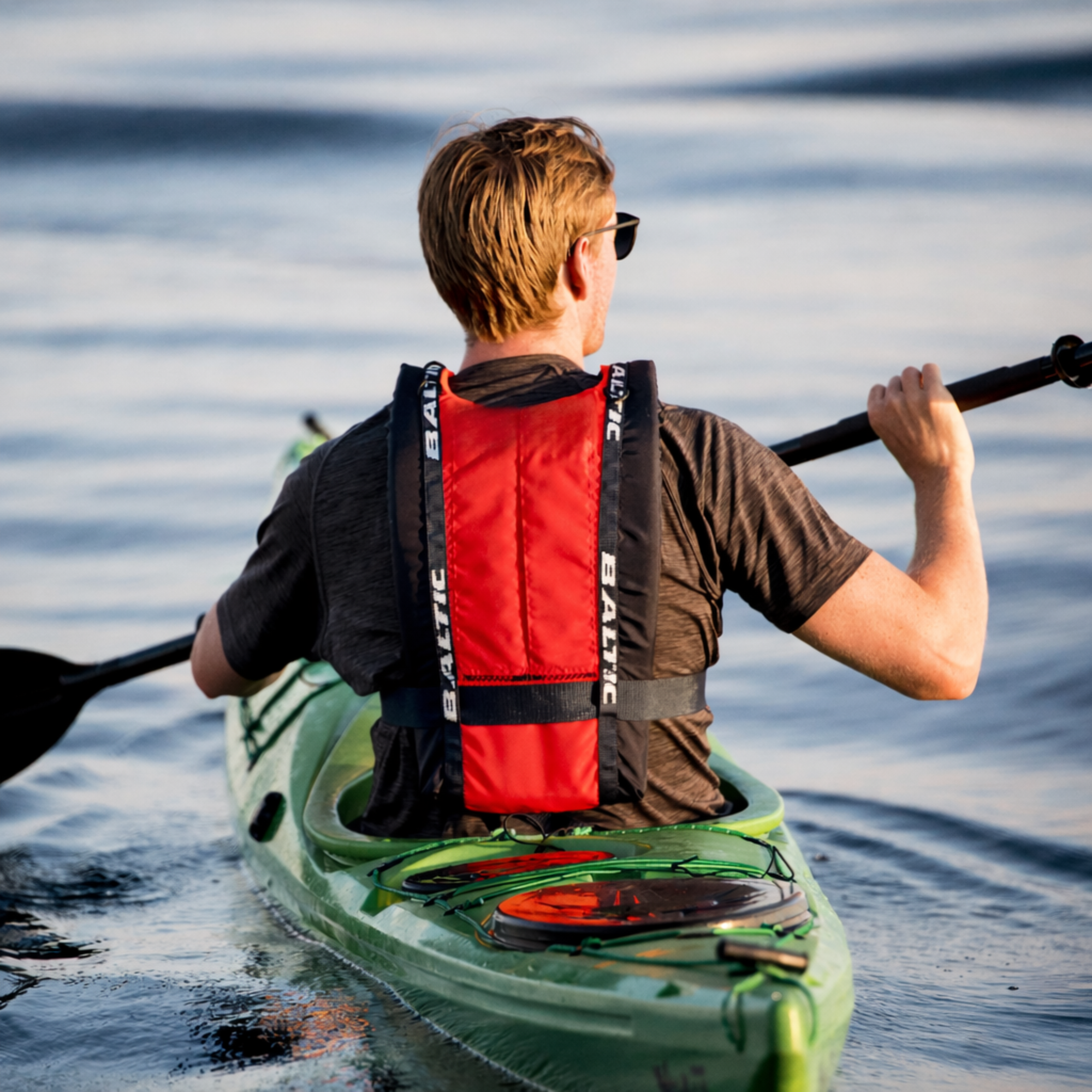 Rear view of kayaker paddling in a green kayak wearing Baltic canoe buoyancy aid showing reinforced back panel, adjustable safety belt, and ergonomic design for comfort and freedom of movement