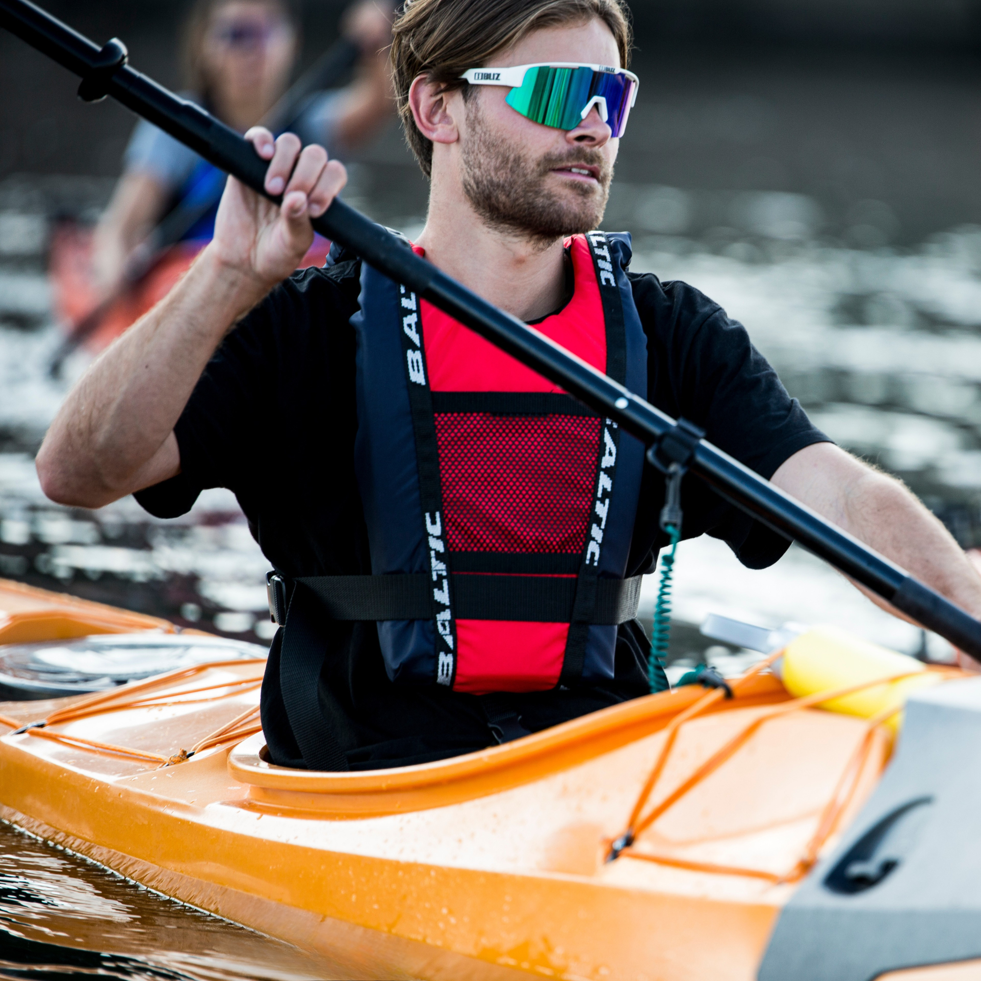 Kayaker wearing Baltic canoe buoyancy aid in red with reinforced shoulder straps, adjustable waist belt, and breathable mesh front pocket while paddling an orange kayak on open water