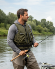 Male angler wearing Baltic Outdoor camo green buoyancy aid with multiple zipped fishing pockets while standing beside a fishing kayak on a calm riverbank