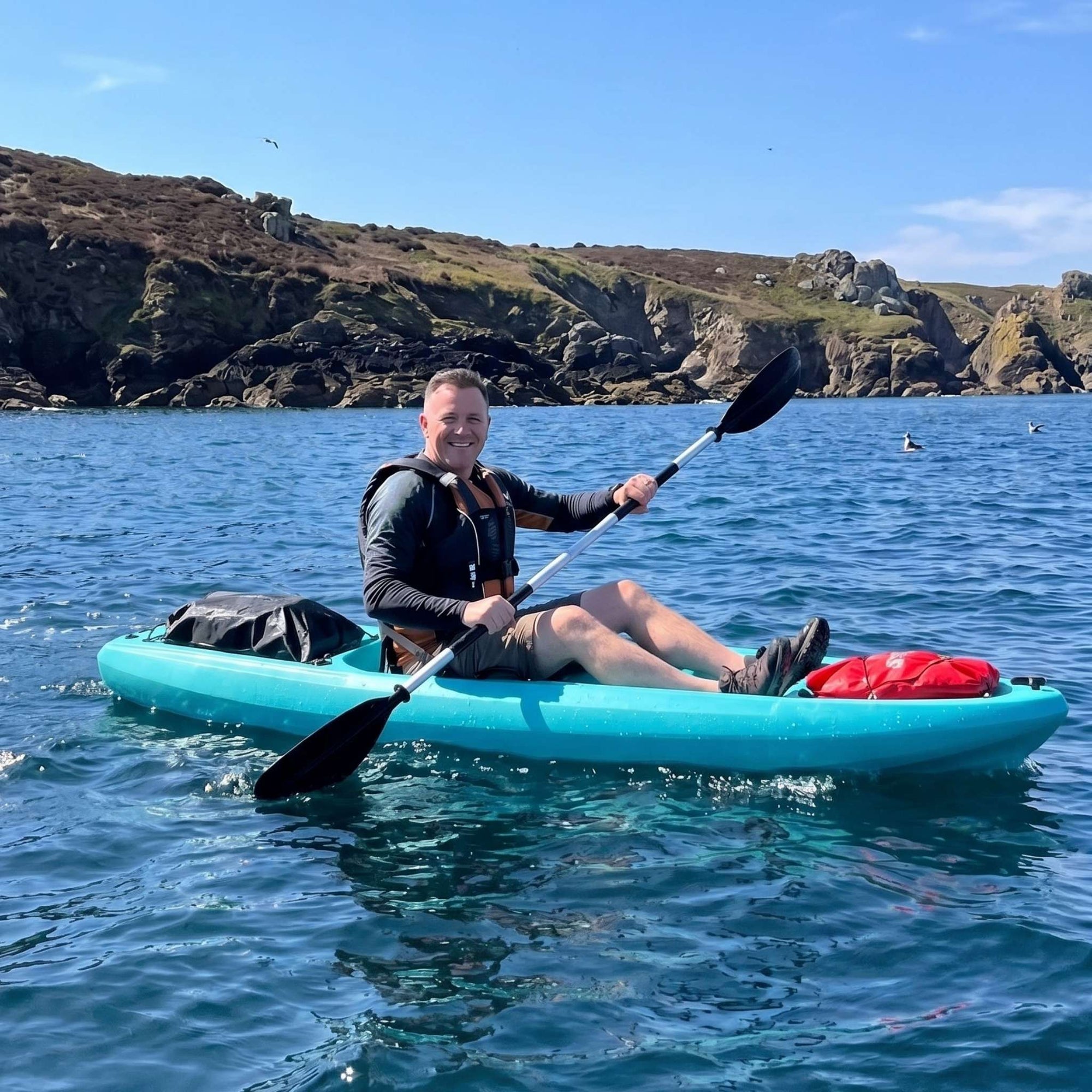 Man paddling single sit on top kayak on clear sea near rocky coastline